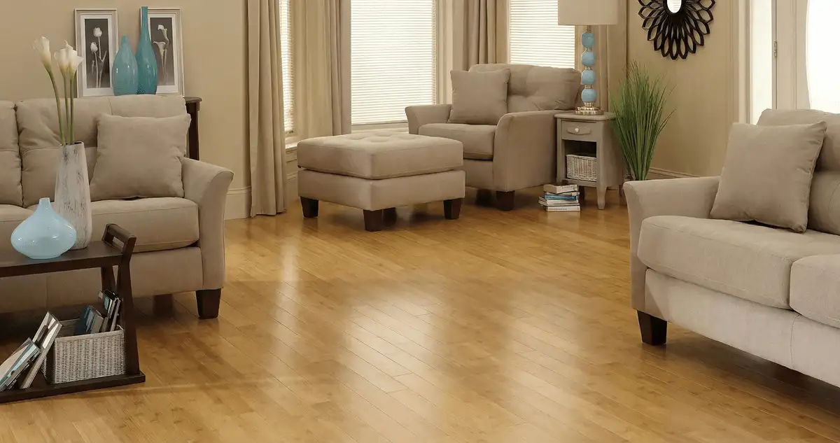 A living room with professionally installed bamboo flooring, beige sofas, a matching ottoman, a side table with a blue lamp, vases, framed pictures, and large windows with blinds and curtains in Santa Ana, CA
