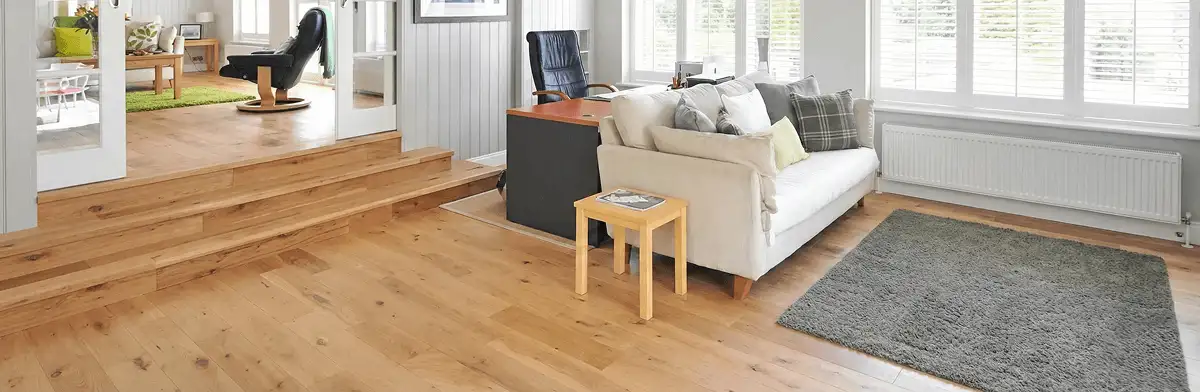 A modern living room with professionally installed laminate flooring, a beige sofa, a small side table, a gray rug, and large windows with white shutters in Santa Ana, CA